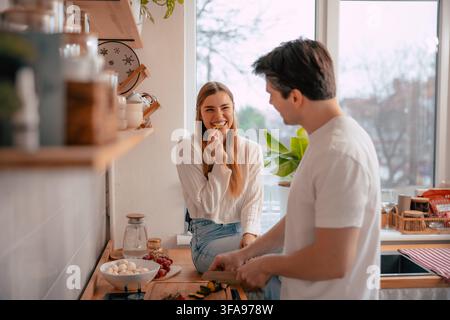 Frau sitzt auf einer Küchenarbeitsplatte und nascht frische Zucchini, während ihr Partner Gemüse für eine gesunde hausgemachte Mahlzeit hackt Stockfoto