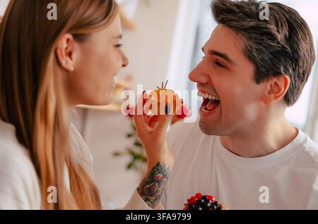 Junge Frau füttert ihren lächelnden Freund verspielt mit einem Obstkuchen, während er ein gemischtes Beerengebäck in ihrer Küchenstube hält Stockfoto