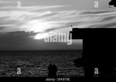 Sonnenuntergang malt den Ozean in warmen orangefarbenen und goldenen Tönen. Silhouette eines Piers und eines Paares auf dem Deck. Das Paar steht nah beieinander und genießt den friedlichen Moment. Perfekt für ro Stockfoto