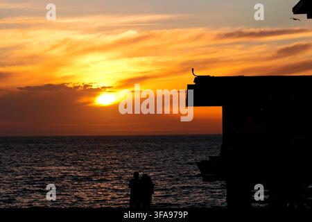 Sonnenuntergang malt den Ozean in warmen orangefarbenen und goldenen Tönen. Silhouette eines Piers und eines Paares auf dem Deck. Das Paar steht nah beieinander und genießt den friedlichen Moment. Perfekt für ro Stockfoto