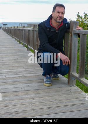 Ein dunkelhaariger, bärtiger, weißer Europäer, der einen Wintermantel trägt, ist entspannt in einem Strandgebiet. Stockfoto
