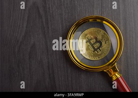 Bitcoin-Münze, die durch eine Lupe auf einem dunklen Holzuntergrund betrachtet wird. Konzept der Kryptowährungsanalyse, digitale Vermögensforschung Stockfoto