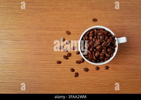 Ansicht von oben einer weißen Tasse, gefüllt mit gerösteten Kaffeebohnen, auf einem Holztisch mit verstreuten Bohnen. Kopierraum für Text. Stockfoto