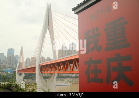 Großes rotes Touristenzeichen mit chinesischen Schriftzeichen neben einer Tagesansicht der Dongshuimen-Brücke, die den Jangtse durchspannt, Chongqing, China. Stockfoto