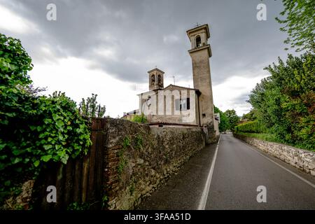 Historische Kirche mit zwei Glockentürmen neben einer schmalen gepflasterten Straße, Steinmauern und dichter grüner Vegetation unter einem dramatischen bewölkten Himmel. Stockfoto