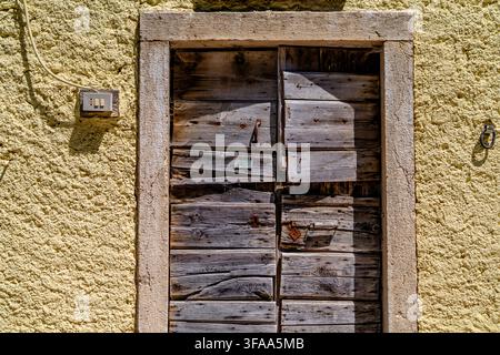 Alte hölzerne Doppeltür aus verwitterten Brettern, die in einen Steinrahmen und eine strukturierte gelbe Putzwand gesetzt ist, mit gealtertem Holz, Nägeln und abgenutzter Oberfläche Stockfoto