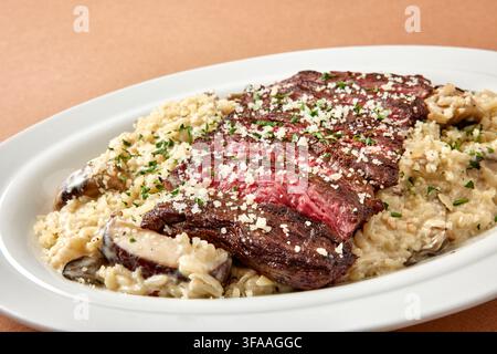 Geschnittenes gegrilltes Rindfleischsteak serviert auf cremigem Risotto mit pochiertem Ei und grüner Zwiebel-Garnitur auf einem weißen Teller vor einem warmen beigen Hintergrund, gehobene Casua Stockfoto