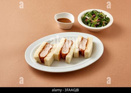 Dickes Katsu-Sandwich im japanischen Stil oder Club-Sandwich mit Schweinekotelett und weichem Brot, serviert auf einem weißen Teller mit Beilagensalat auf einem warmen beigen Hintergrund Stockfoto