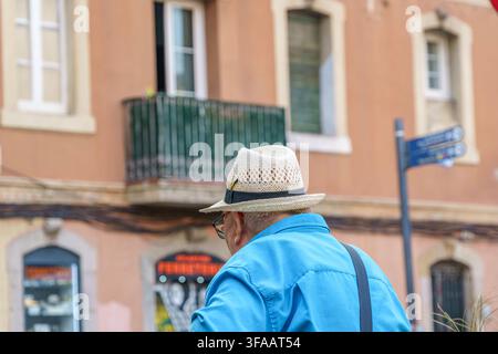 Barcelona – Spanien – 1. August 2026: Älterer Mann mit Strohhut und blauem Hemd geht in einer europäischen Stadtstraße, älterer Tourist, Sommer-Lebensstil Stockfoto