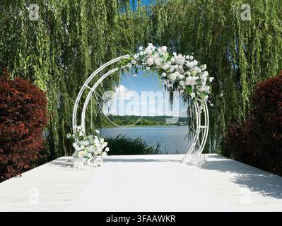Luxuriöse Hochzeitslocation mit romantischem Blumenbogen auf weißem Holzsteg neben einem malerischen See. Eleganter Ort für die Hochzeitszeremonie mit exquisitem Weiß Stockfoto