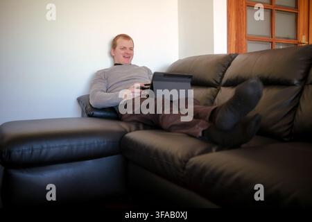 Mann entspannt sich auf einem schwarzen Ledersofa, lächelt und verwendet ein Digitaltablett Stockfoto