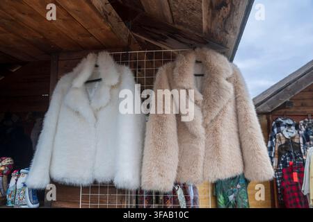 Zwei flauschige Pelzmäntel, einer weiß und einer beige, hängen an einem Drahtgitterregal innerhalb einer rustikalen Holzmarktstandstruktur. Stockfoto