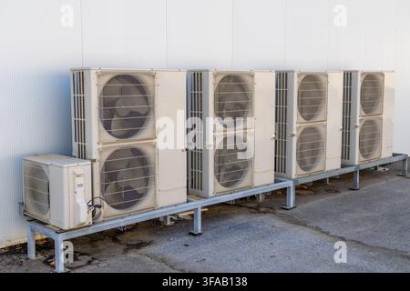 Mehrere beige Außengeräte für Klimaanlagen sind auf einem Metallgestell vor einer strukturierten weißen Wand aufgereiht, was auf eine gewerbliche oder ind Stockfoto