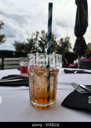 Ein erfrischendes gekühltes Getränk mit einem grün gestreiften Papierstrohhalm, serviert auf einer weißen Tischdecke in einem Terrassenrestaurant im Freien. Stockfoto