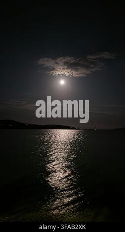 Vollmond scheint über dem ruhigen Meer mit einem dramatischen Lichtreflex auf der Wasseroberfläche und Wolken unter einem dunklen Nachthimmel, Kroatien. Stockfoto