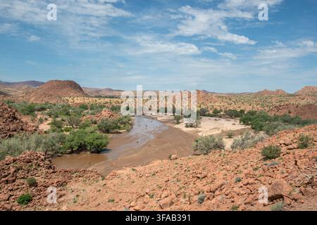 Trüber Fluss windet sich durch eine trockene Wüstenlandschaft in Namibia mit felsigen Hügeln und vereinzelten grünen Bäumen Stockfoto