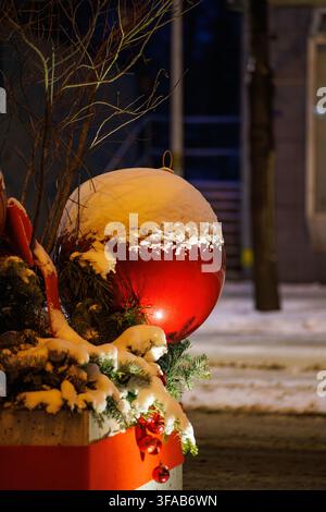 Ein riesiger glänzender roter Weihnachtskugel-Ornamente steht auf immergrünen Kiefernzweigen, die mit frischem Schnee bedeckt sind. Die festliche Straßendekoration wird von warmem Licht beleuchtet Stockfoto