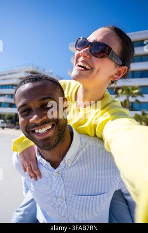 Verspielte Frau reitet auf dem Rücken eines lächelnden Mannes im Freien an einem sonnigen Tag Stockfoto