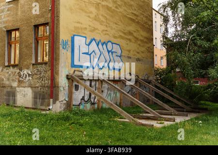 Danzig, Polen - 23. Juli 2026: Graffiti-Tag auf einer texturierten, verwitterten städtischen Gebäudemauer mit blauer Farbe, hölzernen Stützkonstruktionen, Vandalismus, Stadtdesintegration, Untergrundkultur und Grunge-Architektur Stockfoto