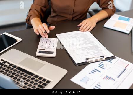 Geschäftsprofi sitzt an einem dunklen Schreibtisch. Hält einen Stift und benutzt einen Taschenrechner, während er Finanzdokumente überprüft. Grafiken. Und ein Laptop. Konzentriert sich auf Aufgaben im Zusammenhang mit Buchhaltung. Budgetierung Stockfoto