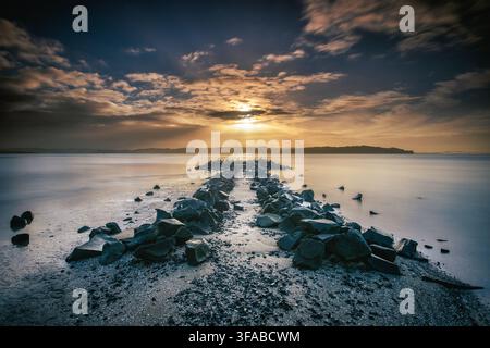Neuseeland - 10. Juni 2021: Blick auf einen felsigen Wellenbrecher, der sich unter einem dramatischen Sonnenuntergangshimmel in das ruhige Meer erstreckt. Stockfoto