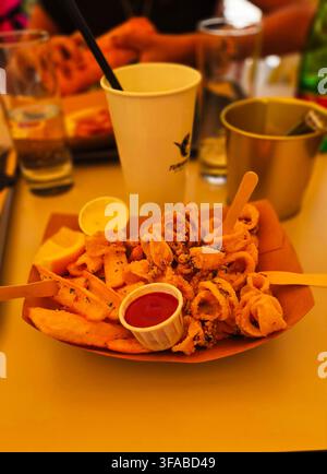Beschriftungen Redaktionell / Beschreibend: Knusprig gebratene Calamariringe und Kartoffelkeile, serviert in einem Kartontray mit Dipsaucen, Zitronenspalter und Getränk Stockfoto
