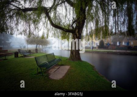Vereinigtes Königreich - 22. April 2023: Blick auf eine Parkszene mit einer Trauerweide, einer grünen Metallbank und einem ruhigen Fluss, der unter einer Steinbrücke fließt Stockfoto