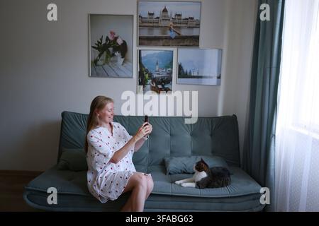 Frau schaut auf Smartphone, während sie neben ihrer Katze auf dem Sofa sitzt, Konzept für Social Media und Freizeit zu Hause. Stockfoto