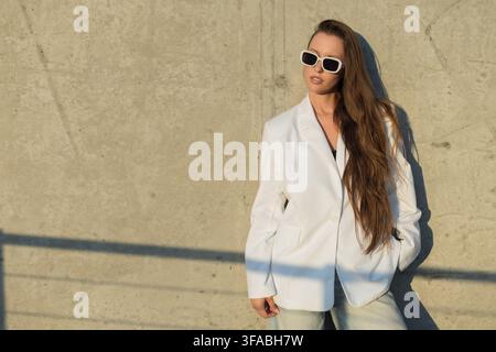 Horizontale Porträt einer stilvollen Frau mit langen Haaren in einem weißen Blazer und einer weißen Sonnenbrille, die an einer Betonwand lehnt. Warmlicht der Goldenen Stunde erzeugt Stockfoto