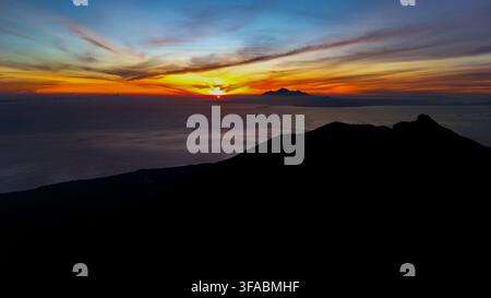 Luftbild des Mount Agung, der sich vor einem lebhaften orangefarbenen und gelben Sonnenuntergangshimmel über dem Meer in Rendang, Bali, Indonesien abhebt Stockfoto