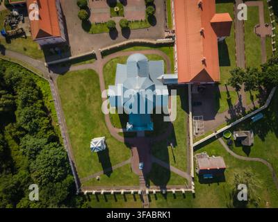 Luftaufnahme der Heiligen-Dreifaltigkeits-Kirche mit ihrem blauen Dach, umgeben von historischen rotgedeckten Gebäuden, grünen Rasenflächen und Steinmauern in Liskiava, Alyt Stockfoto