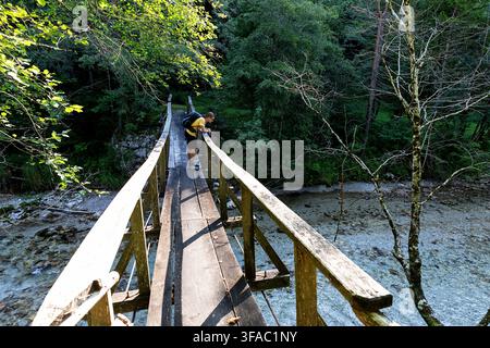 Ein Touristenwanderer steht auf einer hölzernen Fußgängerbrücke über den schönen kristallklaren Savinja-Bergfluss im malerischen Tal von Solcavsko, Slowenien. Stockfoto