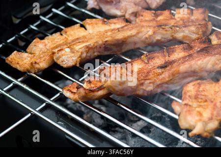 Appetitliche Schweinebauchschnitten, die langsam bis zur Perfektion auf einem Holzkohlegrill geröstet werden, während duftender Rauch von heißen Kohlen im Hinterhof aufsteigt Stockfoto