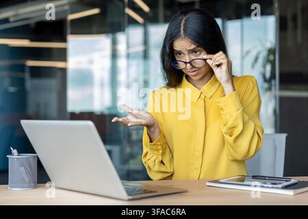 Eine gestresste junge indische Geschäftsfrau schaut auf ihren Laptop-Bildschirm im Büro, senkt ihre Brille mit der Hand und breitet ihre Arme aus. Stockfoto