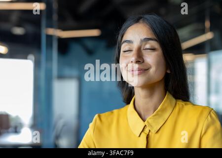 Ruhige und zufriedene junge indische Frau steht im Büro, mit geschlossenen Augen und ruht sich aus. Stockfoto