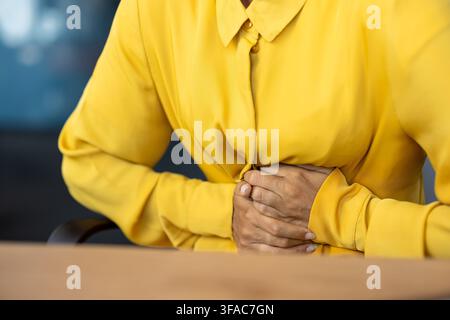 Nahaufnahme eines Körperteils, die Hände einer Frau in einem langen Hemd, die an einem Bürotisch sitzt und ihren Bauch hält, schwere Schmerzen und Unbehagen empfindet Stockfoto