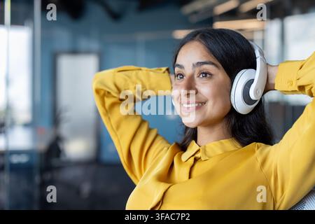 Nahaufnahme einer jungen indischen Frau, die Kopfhörer trägt und in einem Büro sitzt, die Arme hinter dem Kopf verschränkt, lächelnd und entspannt. Stockfoto