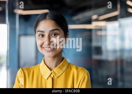 Nahaufnahme-Porträt einer lächelnden jungen indischen Frau in einem gelben Hemd, die in einem Büro steht und in die Kamera schaut. Stockfoto