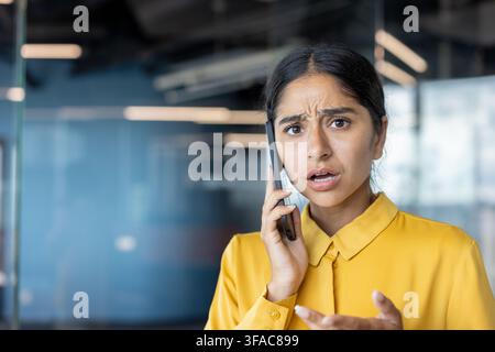 Nahaufnahme-Porträt einer jungen schockierten indischen Frau, die in einem Büro steht, telefoniert und besorgt in die Kamera schaut. Stockfoto