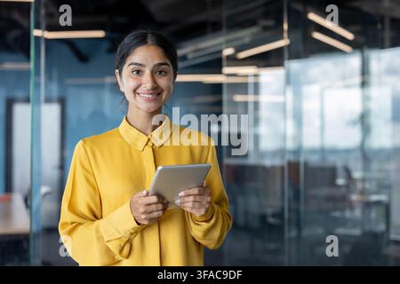 Porträt einer jungen indischen Geschäftsfrau, die im Büro steht, ein Tablet hält und in die Kamera lächelt. Stockfoto