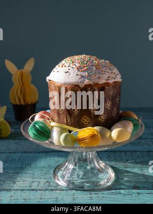 Traditionelle osteuropäische Osterpaska, auch bekannt als Pasca, auf einem gläsernen Kuchenständer präsentiert, umgeben von bunten Dekorationseiern und Frühlingsfeiern Stockfoto