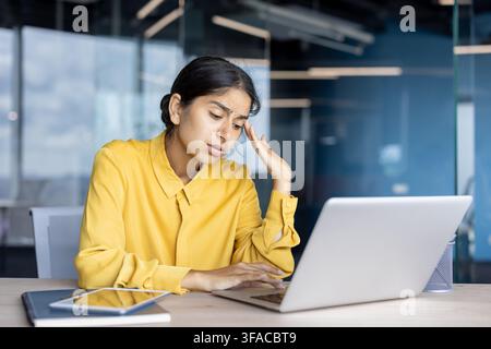 Müde und frustrierte junge indische Frau sitzt am Schreibtisch im Büro vor einem Laptop und hält den Kopf in der Hand. Stockfoto