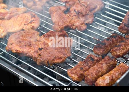 Saftige marinierte Schweinenackensteaks und traditionelle Hackfleischwürste, die über heißen glühenden Holzkohle auf einem Metallgrillrost braten, mit aufsteigendem Rauch Stockfoto
