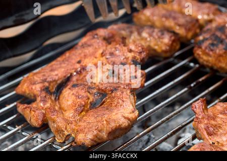 Leckeres gewürztes saftiges Schweinehalssteak und Würste, die über heiß glühender Kohle auf einem Metall-Grillrost für einen schmackhaften rustikalen P zubereitet werden Stockfoto
