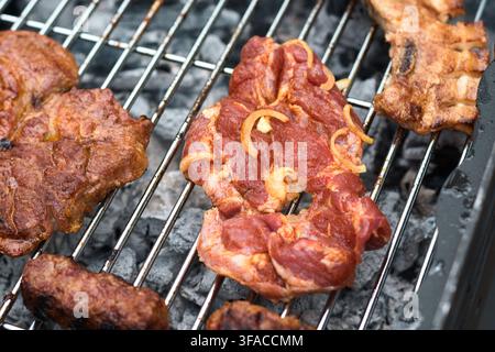 Nahaufnahme eines gewürzten rohen Schweine steaks mit geschnittenen Zwiebeln, der auf einem Metall-Grillrost über glühenden Holzkohlebrocken mit gerösteten Würsten kocht Stockfoto