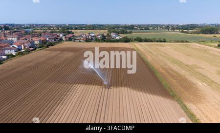 Luftaufnahme eines Bewässerungssprengers, der ein großes braunes Ackerfeld bewässert, mit einer kleinen Stadt im Hintergrund unter einem klaren blauen Himmel in Mediglia, L Stockfoto
