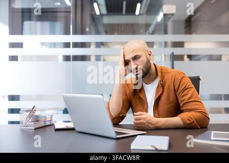 Junger Mann sitzt an seinem Schreibtisch und schaut mit einem gestressten und überforderten Ausdruck auf seinen Laptop-Bildschirm, während er über seine Arbeit nachdenkt. Stockfoto