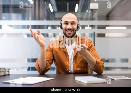 Junger Mann lächelt und gestikuliert mit den Händen während eines Online-Videocalls oder einer Telekonferenz, trägt ein Headset zur Kommunikation in einer modernen Büroumgebung Stockfoto