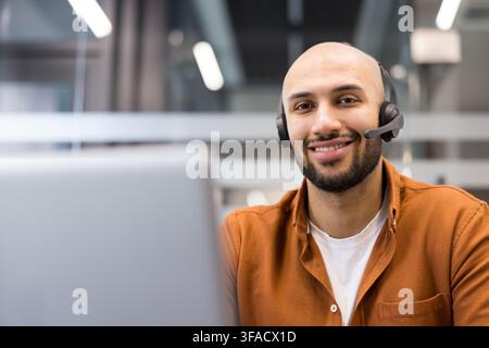 Junger kahlköpfiger Mann mit Bart, der ein Headset trägt und in die Kamera lächelt, der technischen Support für Callcenter und exzellenten Kundenservice von einem Büroarbeitsplatz aus anbietet Stockfoto