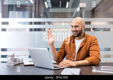 Junger kahlköpfiger Mann mit Bart, der eine Videokonferenz auf einem Laptop abhält, aus einem modernen Büro aus der Ferne arbeitet, lächelt und winkt, online kommuniziert Stockfoto